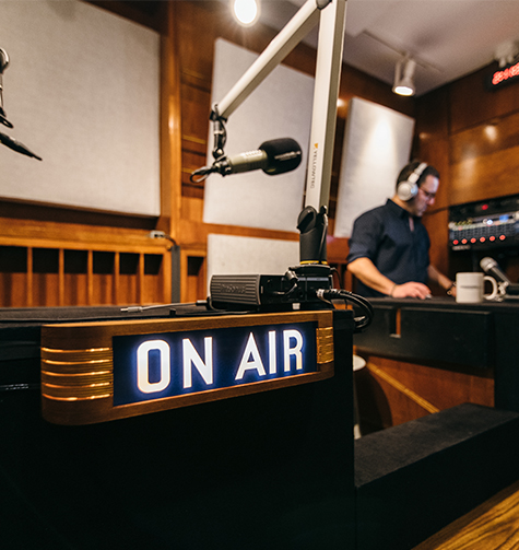 The lit ON AIR sign inside Newsstand Studios, wood paneling and microphone booms behind it